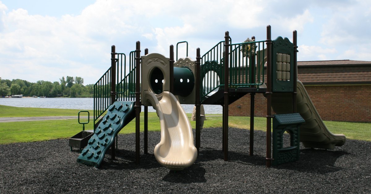 A playground with green slides, tube tunnels, climbing panels, mulch, grass, and a cloudy sky behind it.