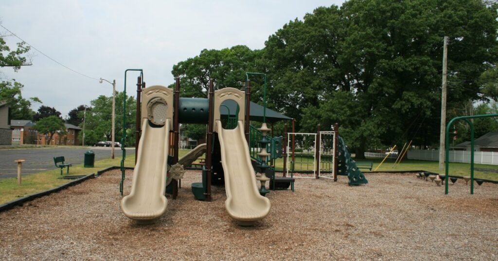 A playground with tan slides, climbing panels, swings, mulch, trees, and a park path under a cloudy sky.