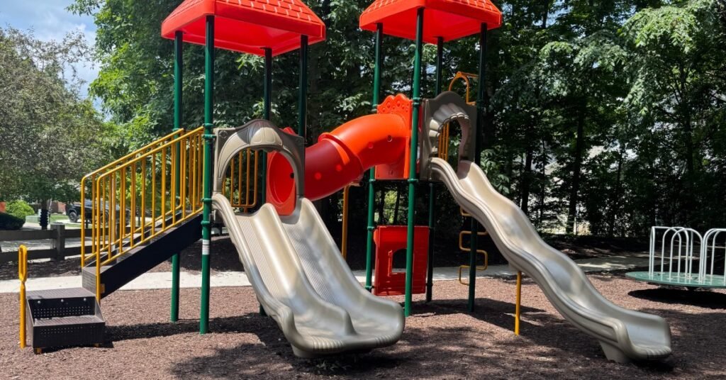 A playground with red tube slides, yellow railings, tan slides, mulch, trees, and bright sunlight overhead.
