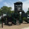 A playground structure with green railings, slides, a tower, trees, and mulch beneath a cloudy blue sky.
