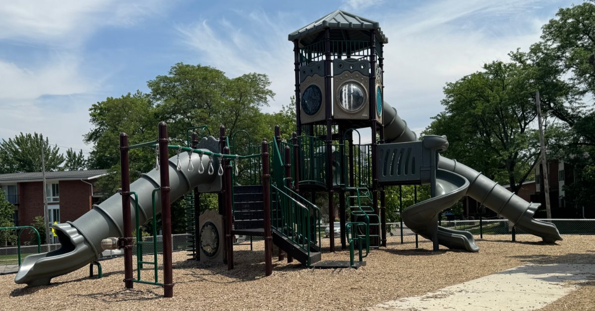 A playground structure with green railings, slides, a tower, trees, and mulch beneath a cloudy blue sky.
