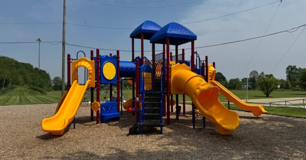 A colorful playground with blue roofs, yellow slides, red posts, mulch, grass, and a road in the background.