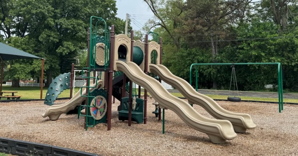 A playground with tan slides, green railings, climbing panels, mulch, trees, and a swing frame nearby.