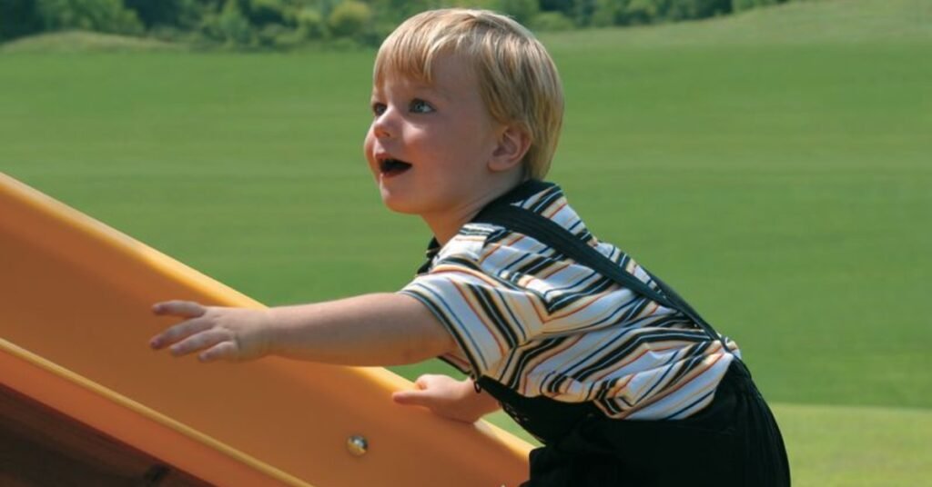 An excited child with a wide smile and curious expression climbing up a yellow portion of a Rainbow playset.