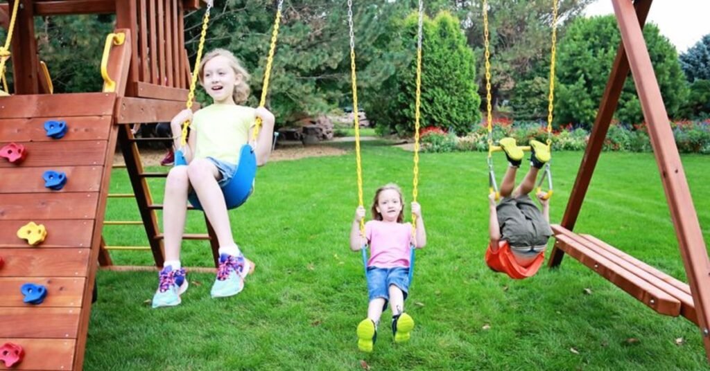 A trio of siblings, all different ages, using the sturdy yellow-and-blue swings of a wooden Rainbow backyard playset.