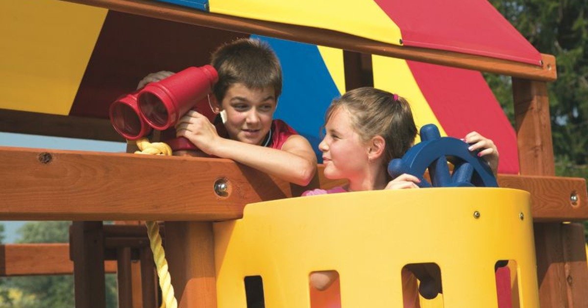 A pair of siblings standing under the shade of a Rainbow playset's colorful canopy and pretending to sail.