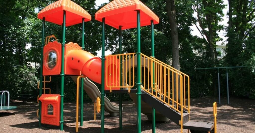 An orange, green, and yellow playset in a partially-shaded area of a public playground, with ladders and slides.