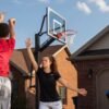 A trio playing basketball in the driveway of a residential house. One person is lifting a hand to block a shot.