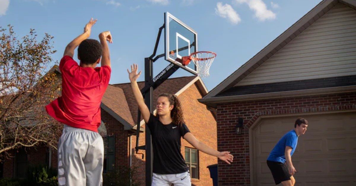 A trio playing basketball in the driveway of a residential house. One person is lifting a hand to block a shot.