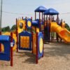 A colorful playground with blue roofs, yellow slides, red posts, wood chips, grass, trees, and open sky.