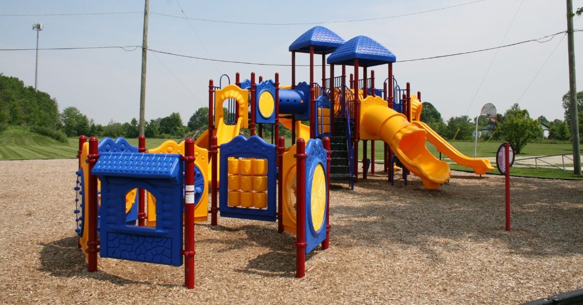 A colorful playground with blue roofs, yellow slides, red posts, wood chips, grass, trees, and open sky.