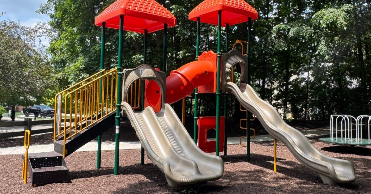 A playground with red roofed towers, cream slides, green posts, a red tunnel, steps, railings, trees, and mulch.