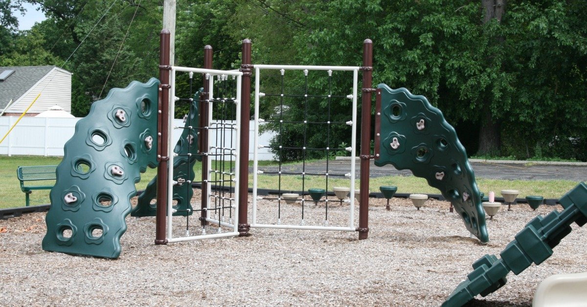 A playground climbing area with green panels, rope nets, brown posts, stepping pods, trees, a fence, and mulch.