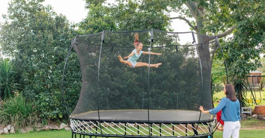 A girl jumping on a large backyard trampoline with black safety netting while a woman stands nearby on grass.