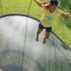 A girl jumping on a round backyard trampoline with black safety netting, surrounded by green grass in sunlight.