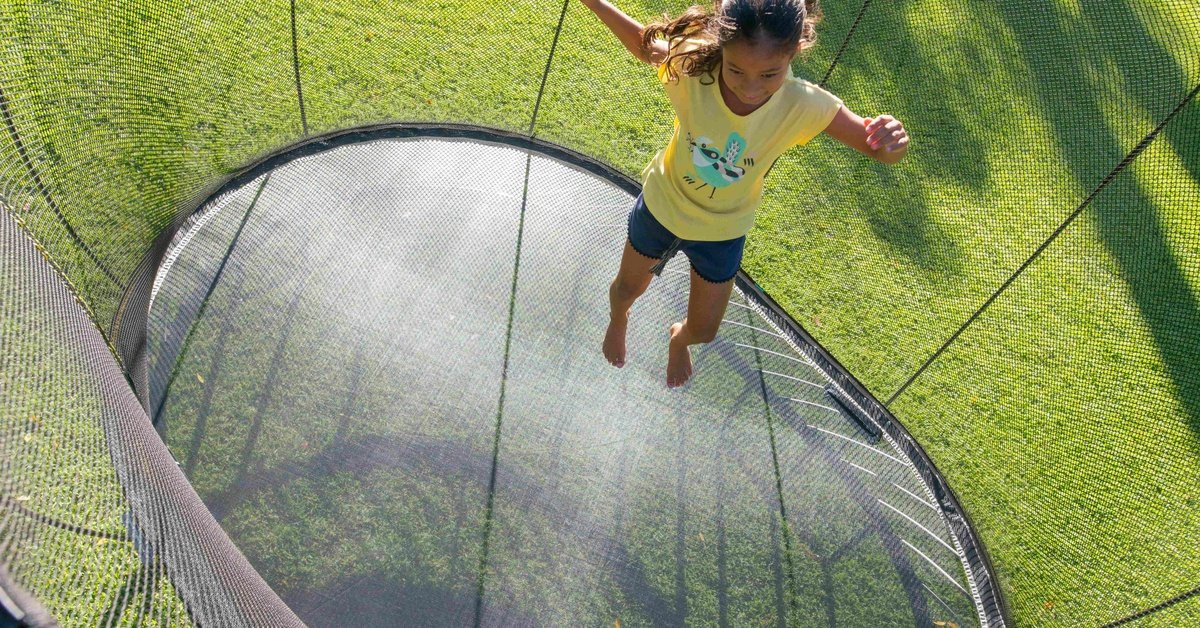A girl jumping on a round backyard trampoline with black safety netting, surrounded by green grass in sunlight.