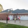 A brick family home with three kids playing basketball and guardians sitting in folding chairs spectating.