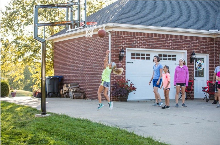 One girl at home in the driveway shooting a layup with a basketball with three people spectating behind her.
