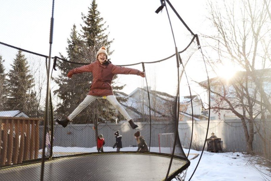 A young girl bounces on her Springfree trampoline on a cold winter day. There is snow on the ground and she wears a coat.