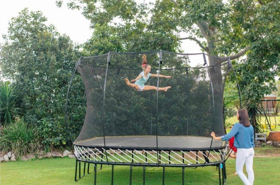 A young girl doing cheer jumps on a Springfree trampoline in the backyard. Mom watches on the side of the net.