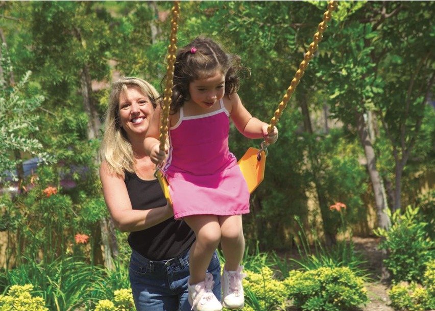 A mom smiles as she pushes her young daughter on the swing in the backyard. The daughter wears a pink dress.