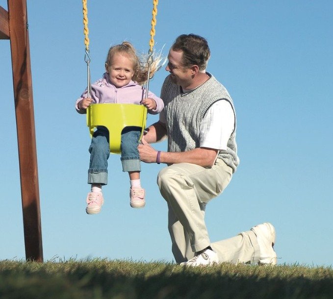 A father kneels next to his young daughter on her baby swing. The swing is yellow and it's sunny outside.