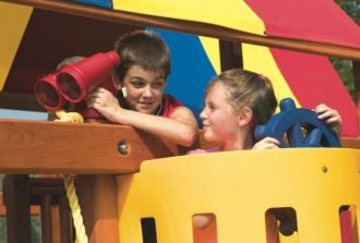 A pair of siblings standing under the shade of a Rainbow playset's colorful canopy and pretending to sail.