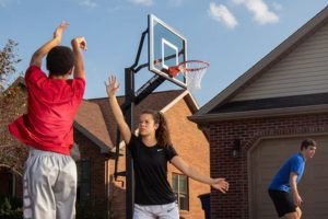 A trio playing basketball in the driveway of a residential house. One person is lifting a hand to block a shot.