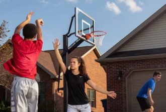 A trio playing basketball in the driveway of a residential house. One person is lifting a hand to block a shot.