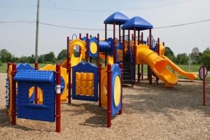 A colorful playground with blue roofs, yellow slides, red posts, wood chips, grass, trees, and open sky.