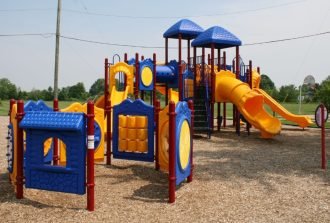 A colorful playground with blue roofs, yellow slides, red posts, wood chips, grass, trees, and open sky.