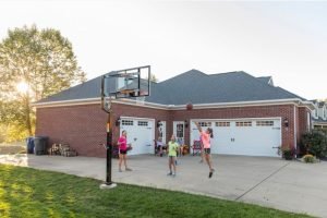 A brick family home with three kids playing basketball and guardians sitting in folding chairs spectating.