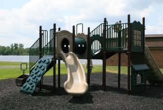 A playground with green slides, tube tunnels, climbing panels, mulch, grass, and a cloudy sky behind it.