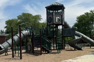 A playground structure with green railings, slides, a tower, trees, and mulch beneath a cloudy blue sky.