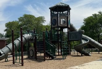 A playground structure with green railings, slides, a tower, trees, and mulch beneath a cloudy blue sky.