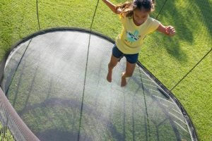 A girl jumping on a round backyard trampoline with black safety netting, surrounded by green grass in sunlight.